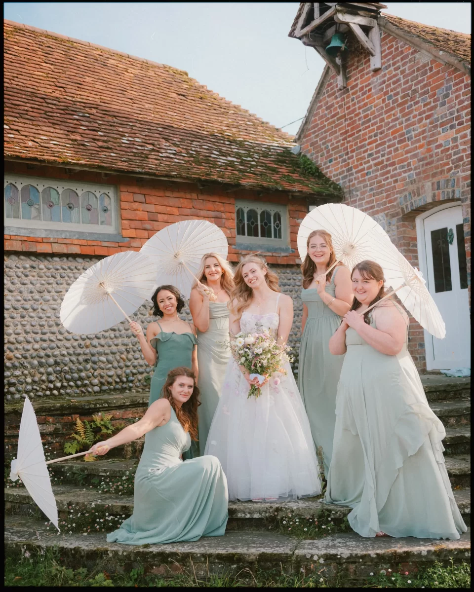 A bride in a white dress stands on stone steps, surrounded by five bridesmaids in sage green with white parasols, in front of a rustic brick building—capturing the charm of a Montague Farm Wedding and festival-style Sussex weddings.