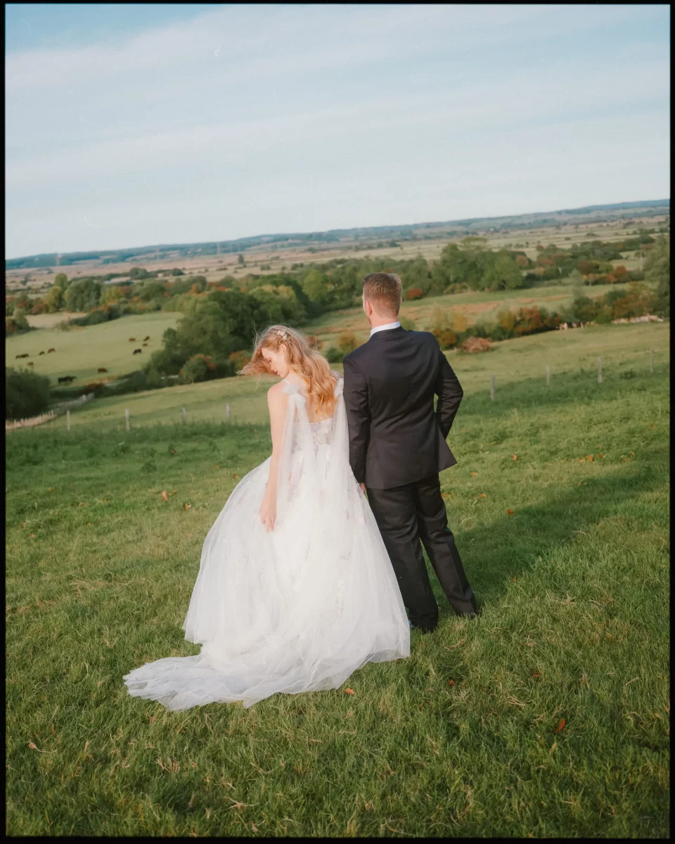 A bride in a white gown and a groom in a black suit stand on a grassy hill at Montague Farm, looking out over a scenic countryside—perfect for festival-style Sussex weddings under a clear sky.