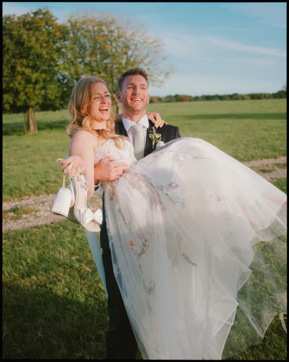 A groom in a suit joyfully lifts his smiling bride, who holds her shoes and wears a flowing white dress, in a sunlit outdoor setting at Montague Farm—perfect for festival-style Sussex weddings amid green grass and trees.
