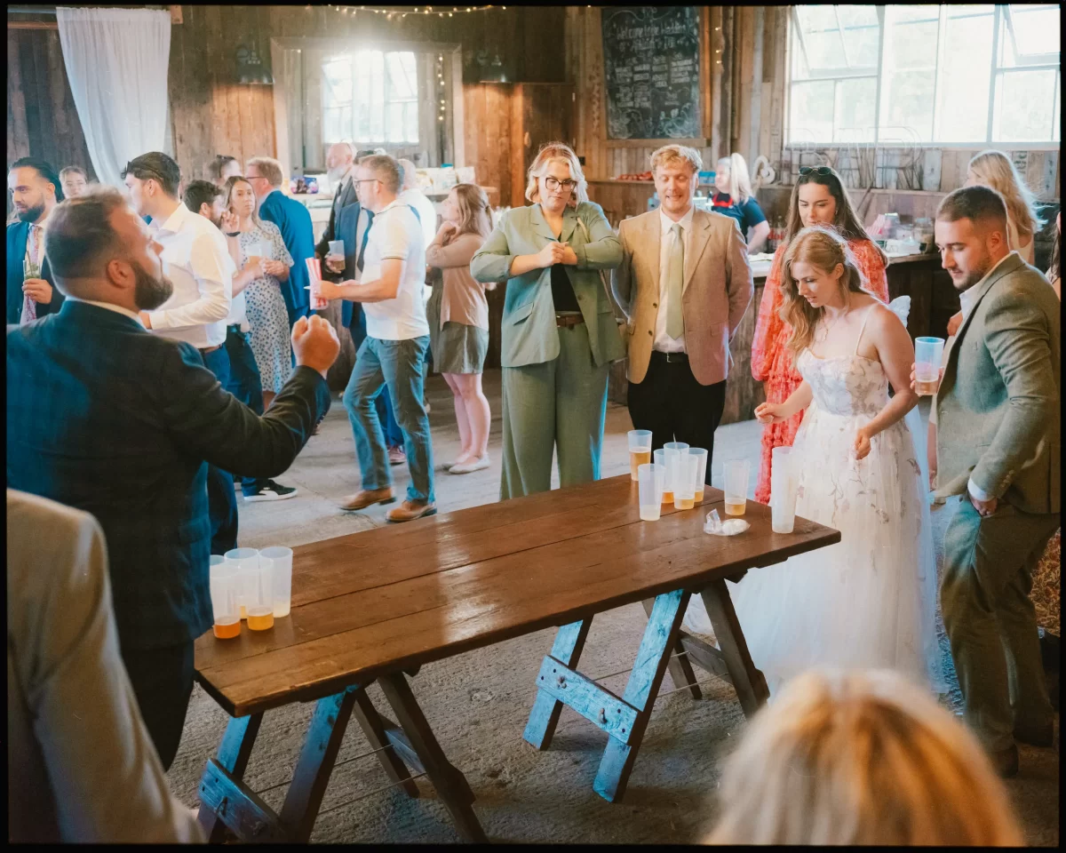 At a Montague Farm Wedding, the bride and guests in formal attire play beer pong on a wooden table inside a rustic venue with wooden walls and large windows, creating a lively festival-style Sussex weddings atmosphere.
