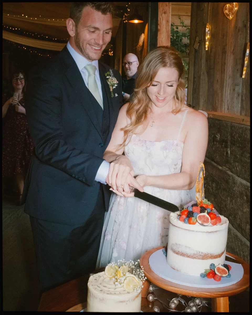 A smiling couple in formal attire cuts a white, berry-topped cake at their Montague Farm wedding, a festival-style Sussex celebration. Another decorated cake sits nearby as string lights twinkle in the rustic indoor venue.