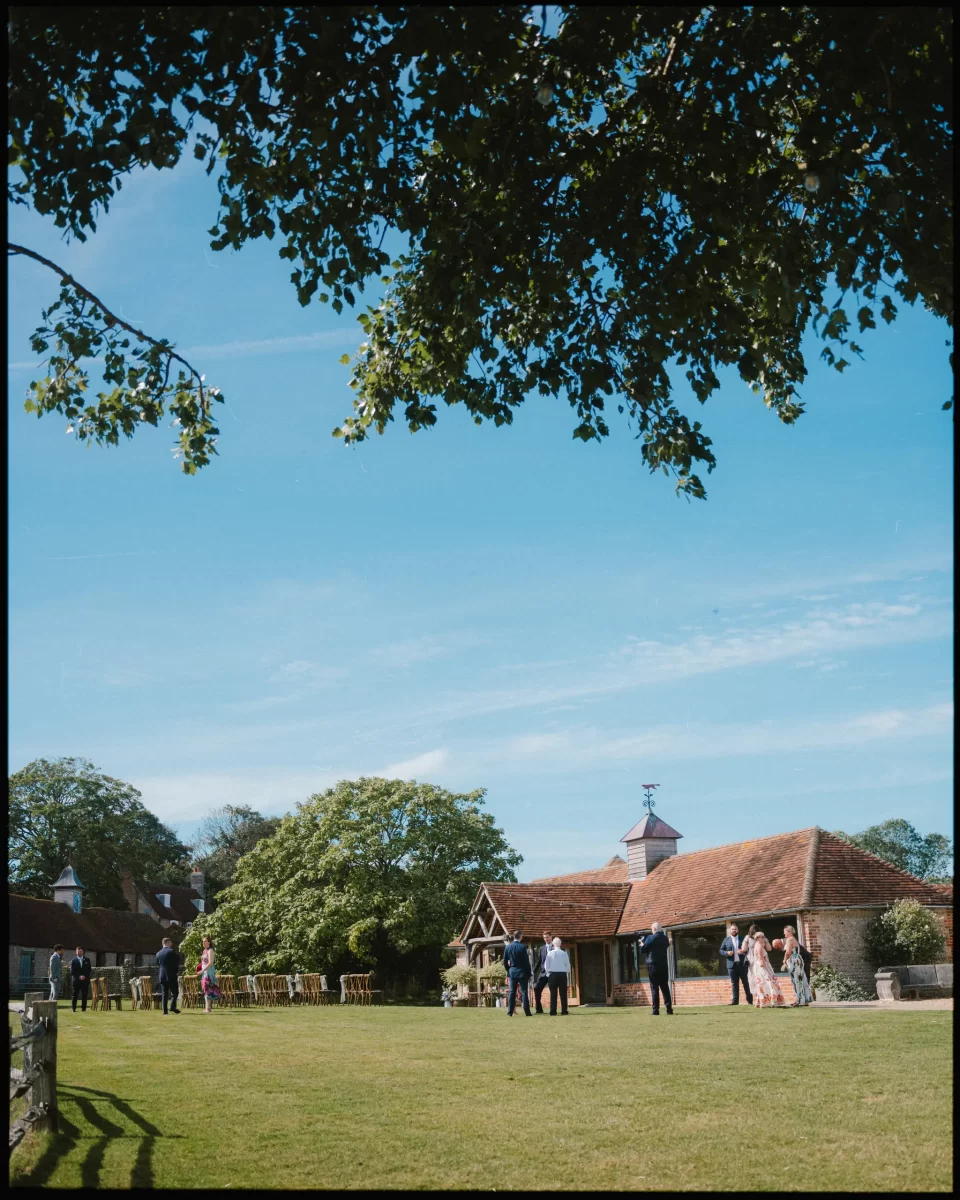 A group of people stand and chat on a large grassy lawn under a sunny blue sky at Montague Farm, perfect for festival-style Sussex weddings, with trees, a wooden fence, and a rustic building with a tiled roof in the background.