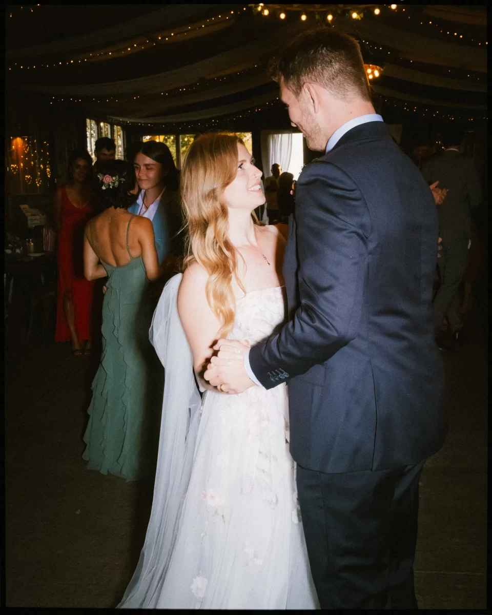 A bride and groom share a first dance at their Montague Farm wedding reception, smiling at each other under string lights. Guests and bridesmaids in dresses complete this festival-style Sussex wedding celebration.