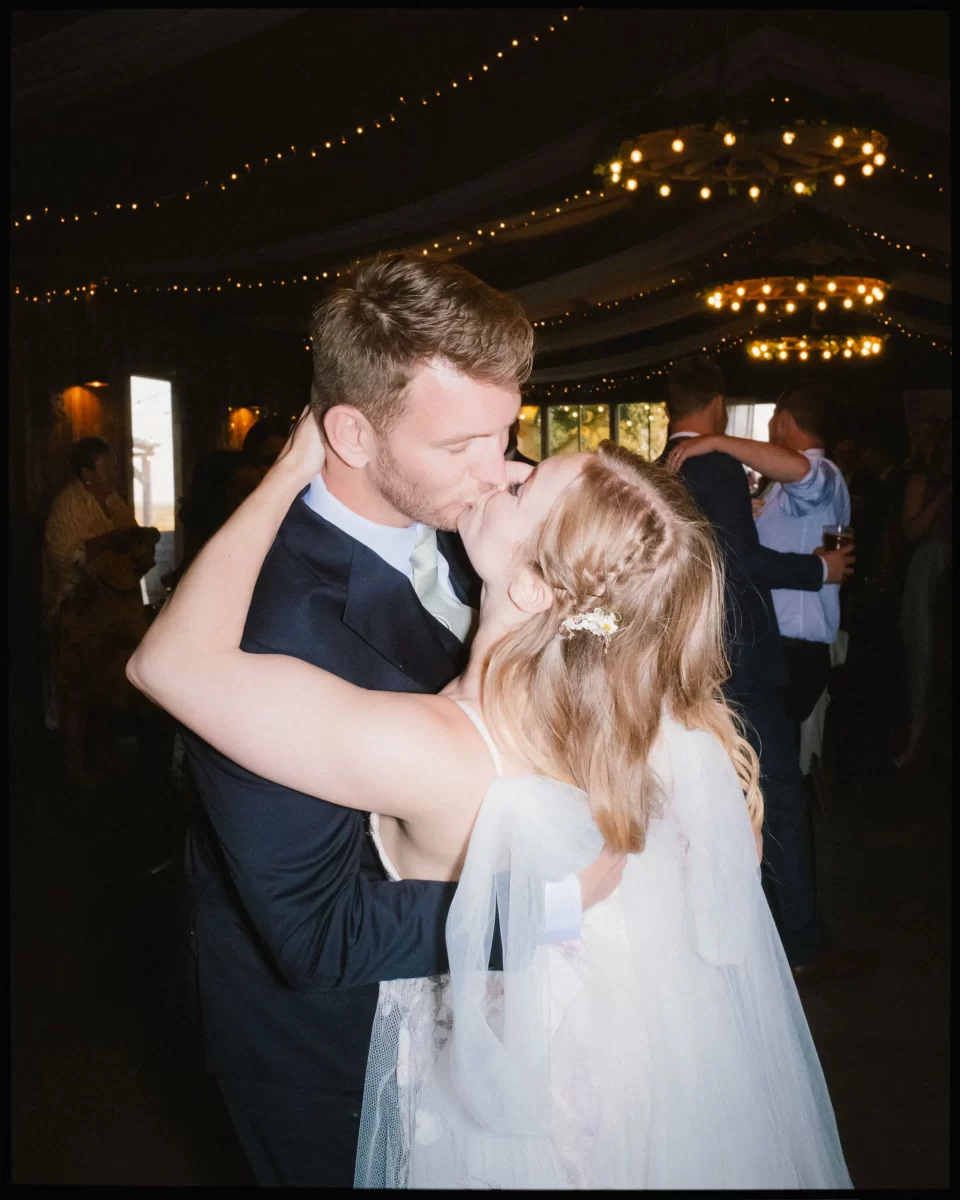 A bride and groom share a kiss while dancing at their Montague Farm wedding reception, surrounded by guests and warm string lights overhead, capturing the essence of festival-style Sussex weddings.