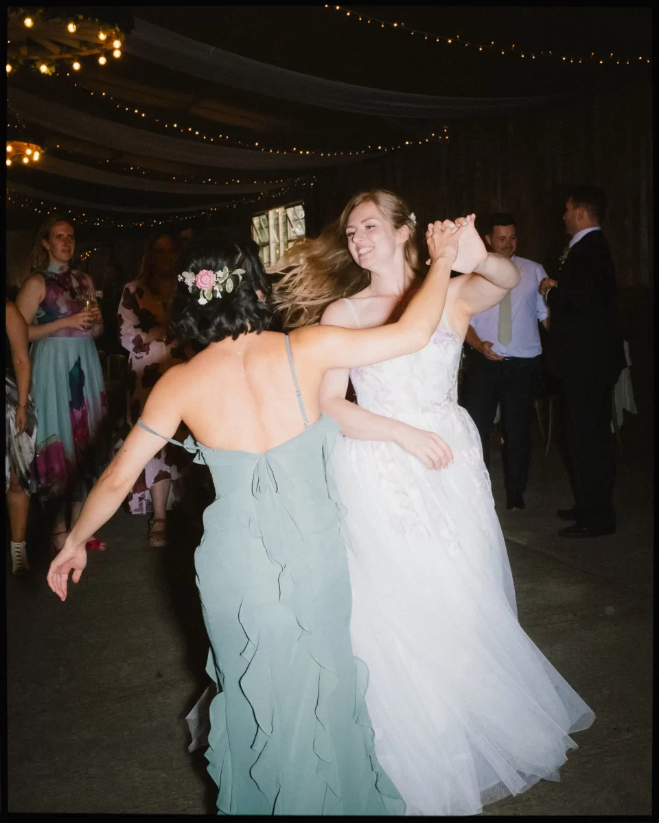 Two women in formal dresses, one in a wedding gown and the other in sage green with flowers in her hair, joyfully dance together at a Montague Farm Wedding, celebrating festival-style Sussex weddings under twinkling string lights.