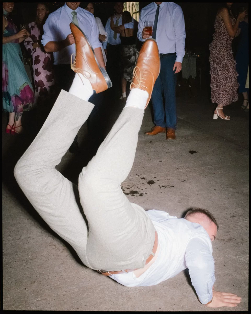 A man in dress clothes breakdances on the floor at a lively Montague Farm Wedding, surrounded by people watching and smiling, capturing the fun spirit of festival-style Sussex weddings.