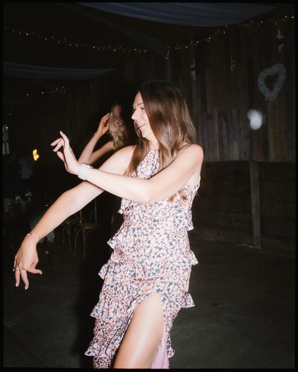 A woman in a pink floral dress dances joyfully in a dimly lit room with wooden walls and string lights at a Montague Farm Wedding, while another woman smiles in the background, capturing the festival-style Sussex weddings vibe.