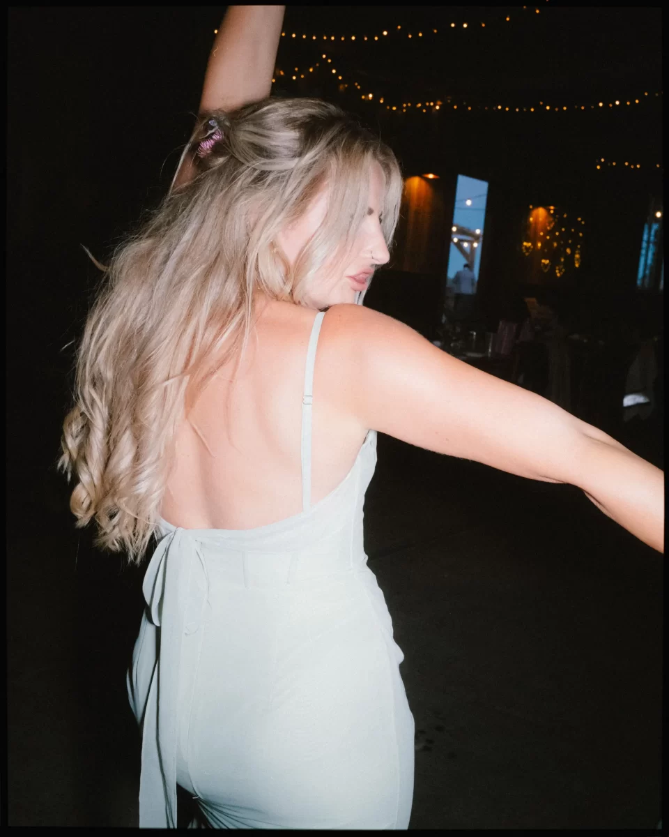 A woman with long blonde hair in a light dress dances with one arm raised, surrounded by dim lighting and string lights—a scene reminiscent of a Montague Farm Wedding or festival-style Sussex weddings.