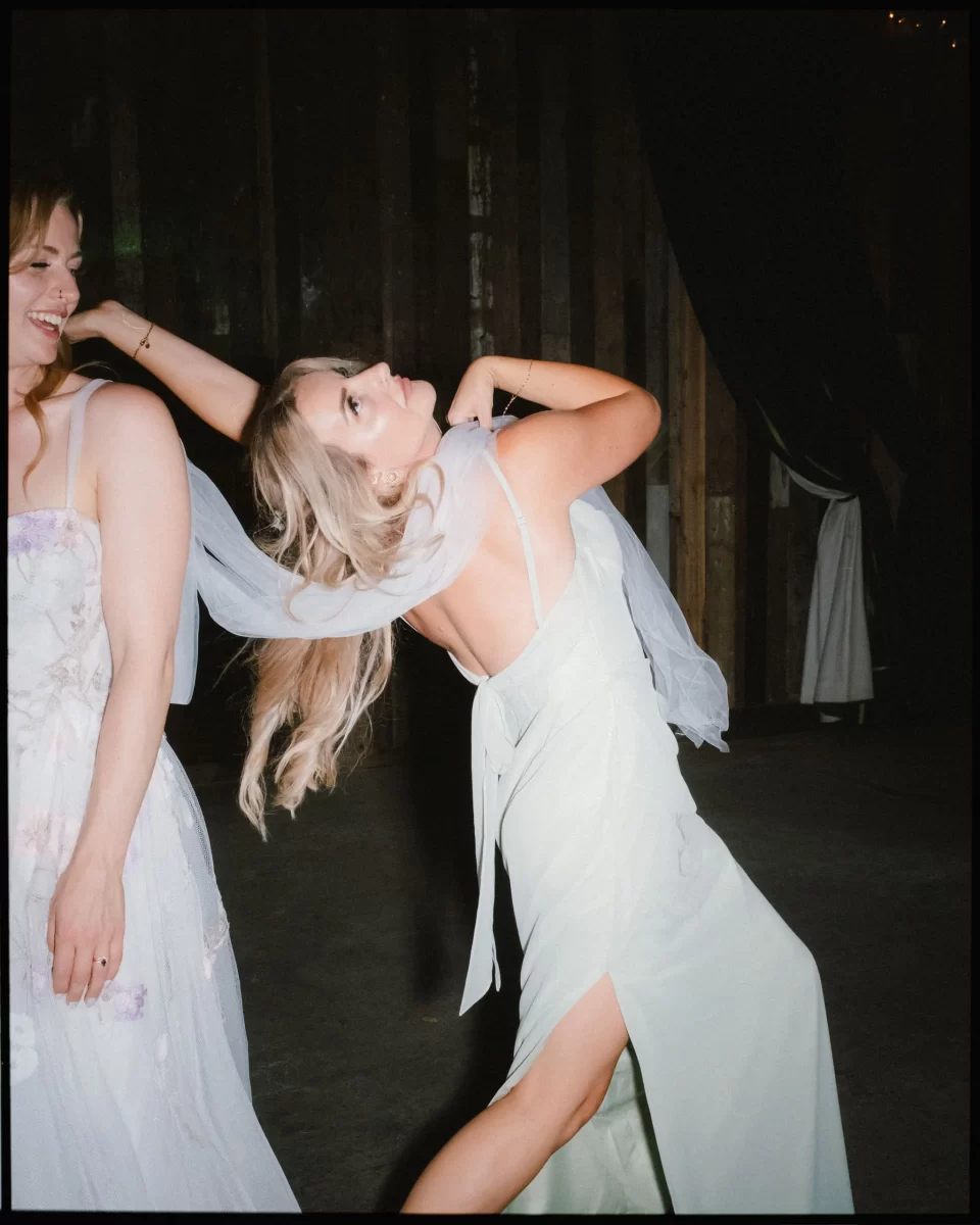 Two women in light-colored dresses dance energetically indoors at a Montague Farm Wedding; one’s veil flies as she leans back dramatically, her hair swinging, while the other smiles, partially out of frame—capturing that festival-style Sussex weddings vibe.