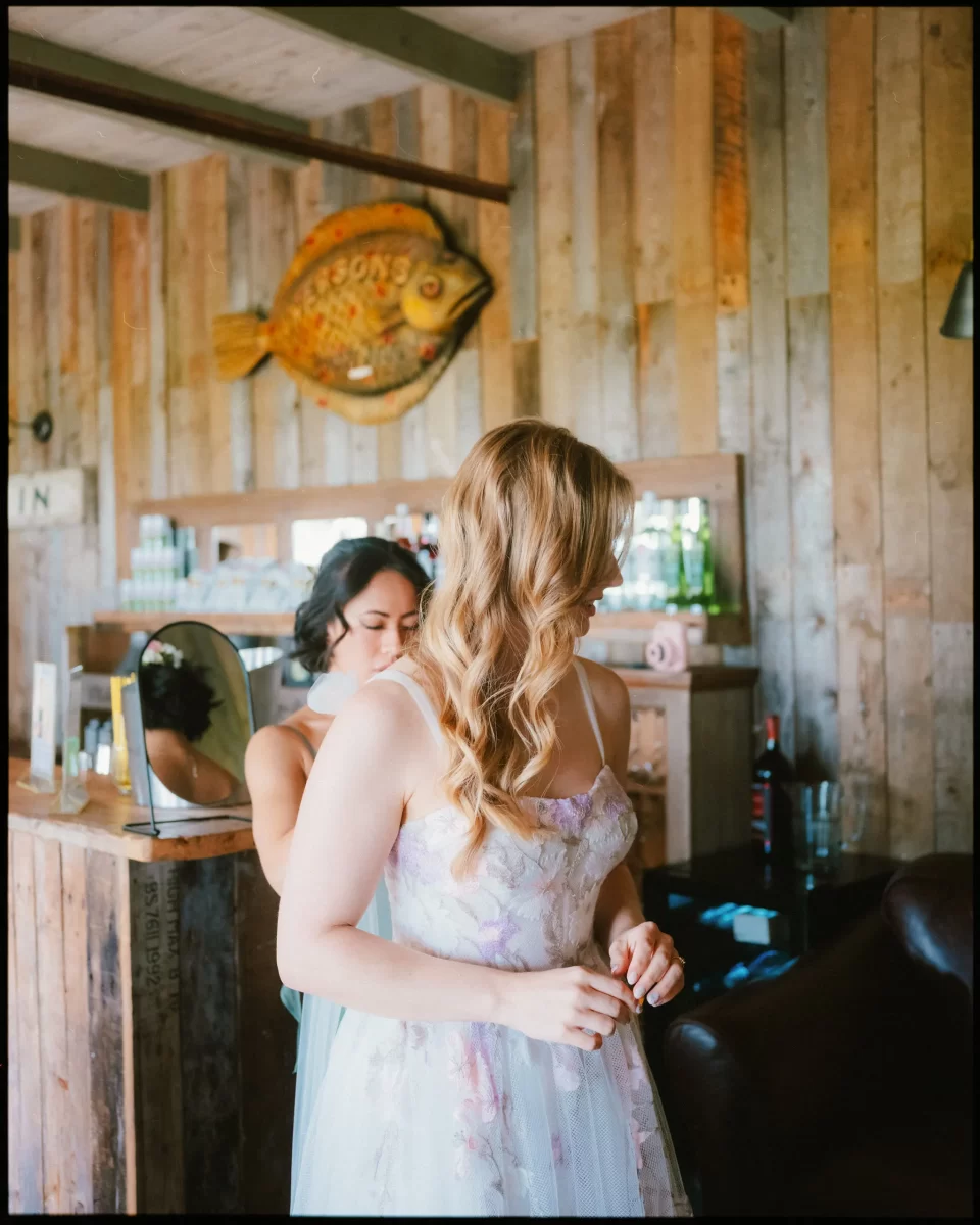 A bride in a floral dress stands indoors as another helps her from behind at a Montague Farm Wedding. The rustic bar, wooden walls, and large fish decoration evoke the charm of festival-style Sussex weddings.