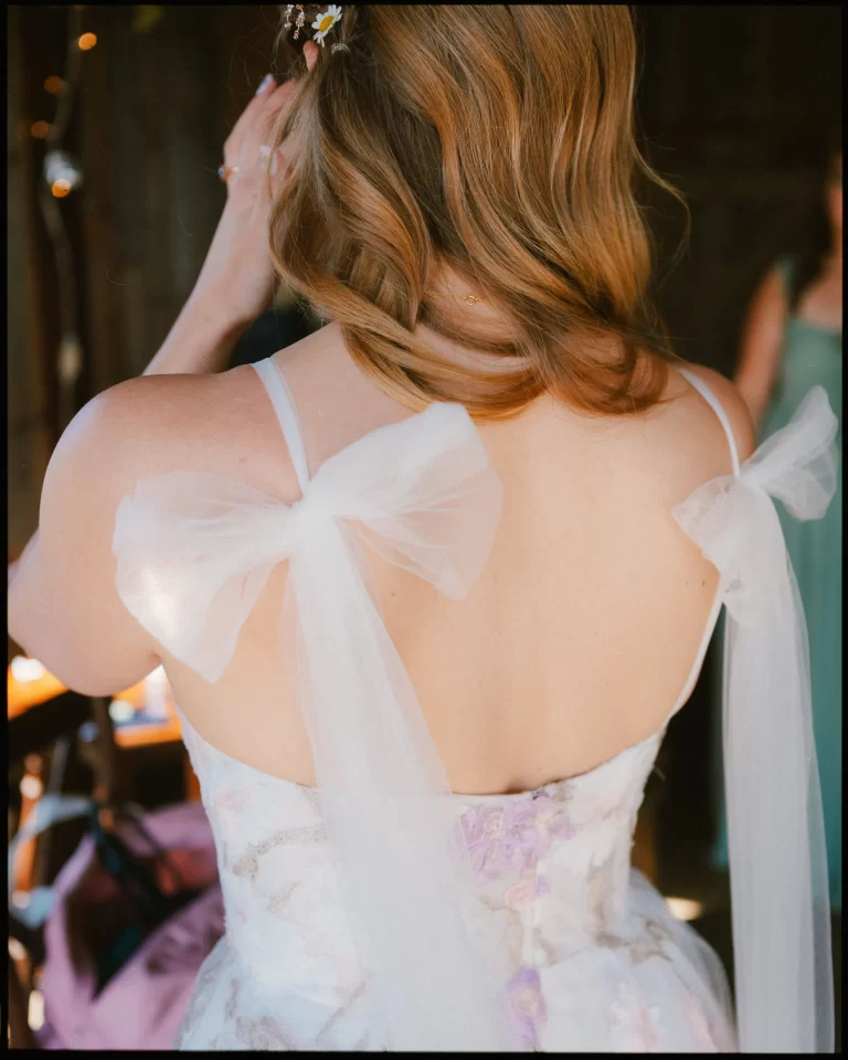 A bride with wavy, light brown hair is seen from behind at a Montague Farm Wedding, wearing a dress with sheer white ribbon bows on the straps. She touches her face as soft sunlight highlights her back—a perfect moment at a festival-style Sussex wedding.