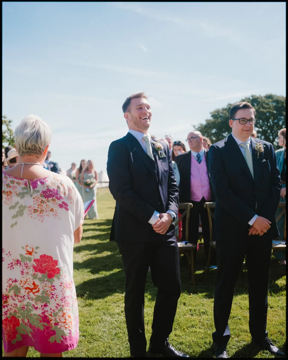 At a Montague Farm Wedding, a groom in a black suit stands outdoors, smiling and laughing with another suited guest beside him, as guests enjoy the festival-style Sussex wedding ceremony. A woman in a floral dress stands in the foreground.