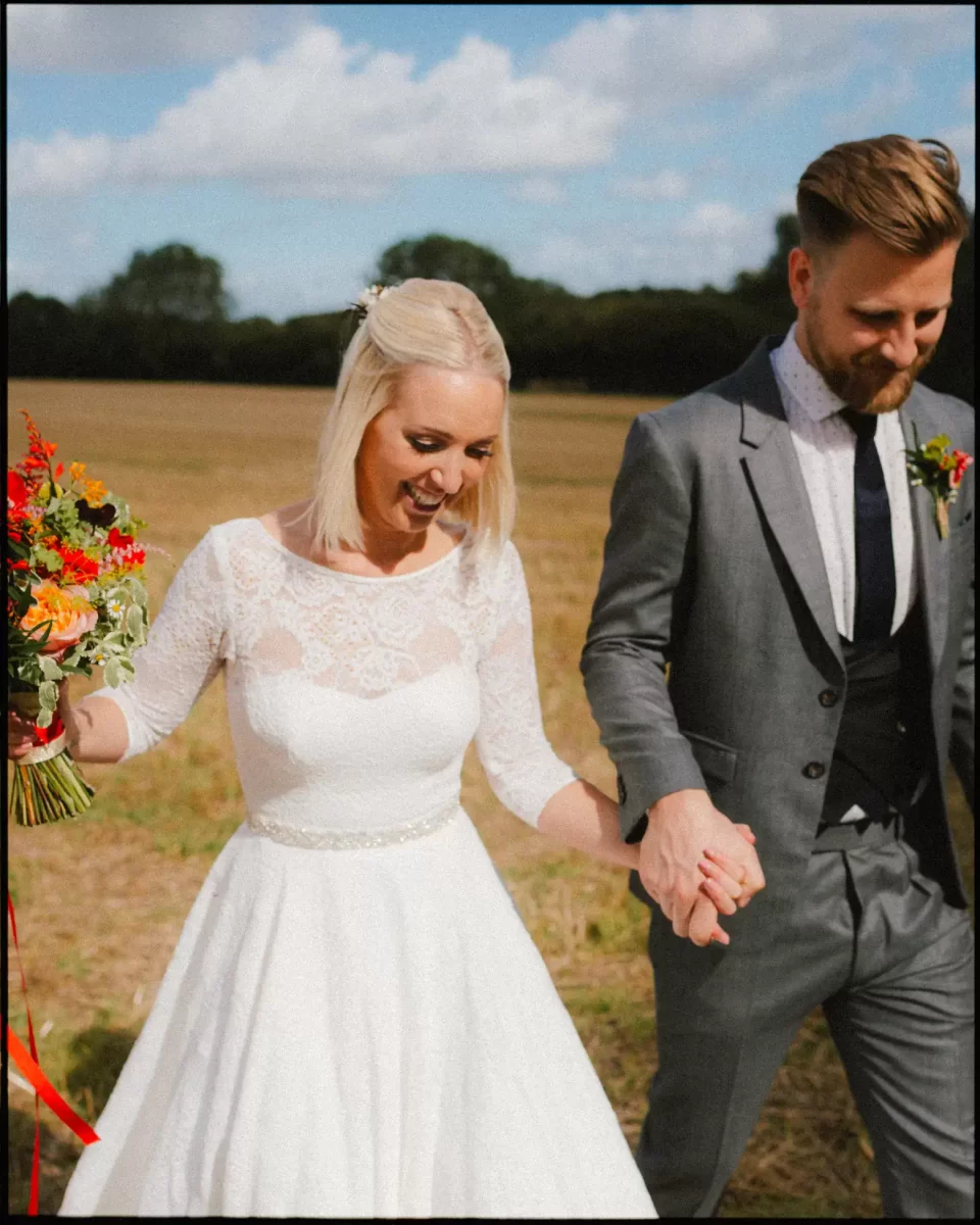 bride and groom walking through field smiling in the sunshine near the bell in ticehurst