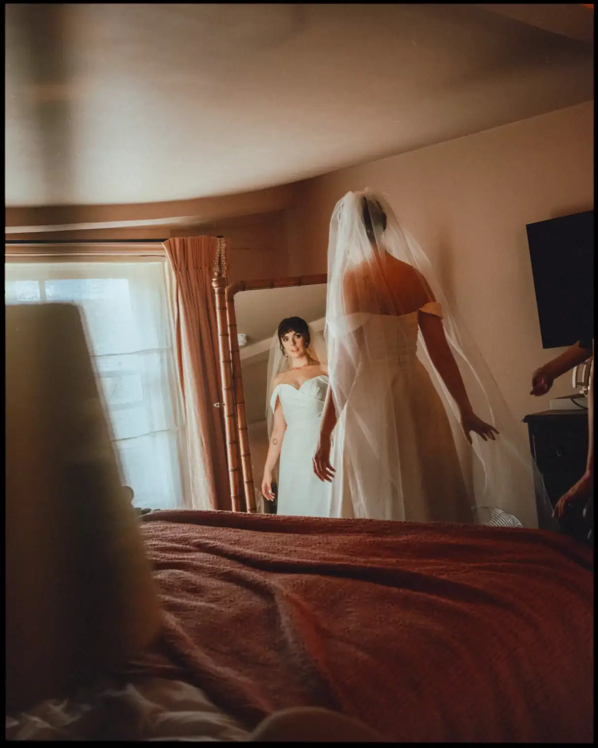 A bride in an off-the-shoulder wedding dress and veil stands before a mirror, gazing at her reflection in a softly lit room—a perfect moment captured by a Rye wedding photographer at the charming George Hotel Rye.
