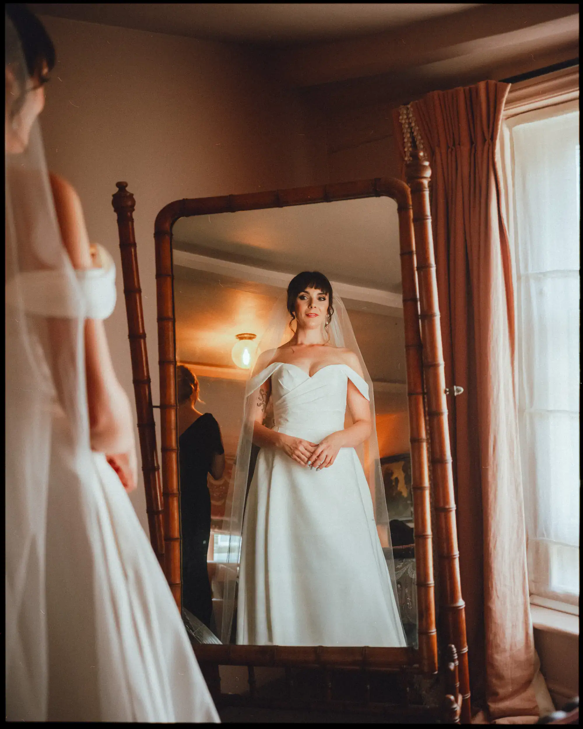 A bride in an off-the-shoulder white gown and veil stands before a mirror at an East Sussex wedding venue, warm light and peach curtains casting soft shadows—a perfect moment for a Rye wedding photographer to capture.