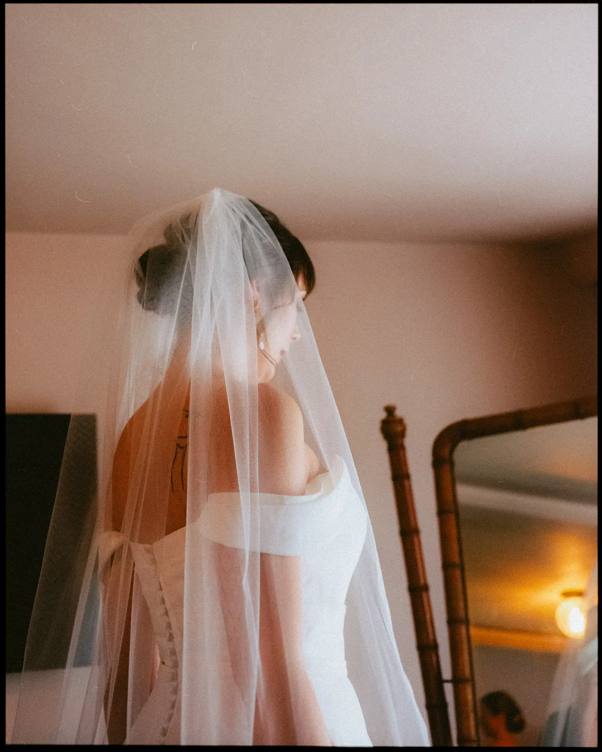 A bride in an off-shoulder white wedding dress and veil stands with her back to the camera, looking toward a mirror in a softly lit room—perfect for candid wedding photography Rye at the George Hotel Rye.