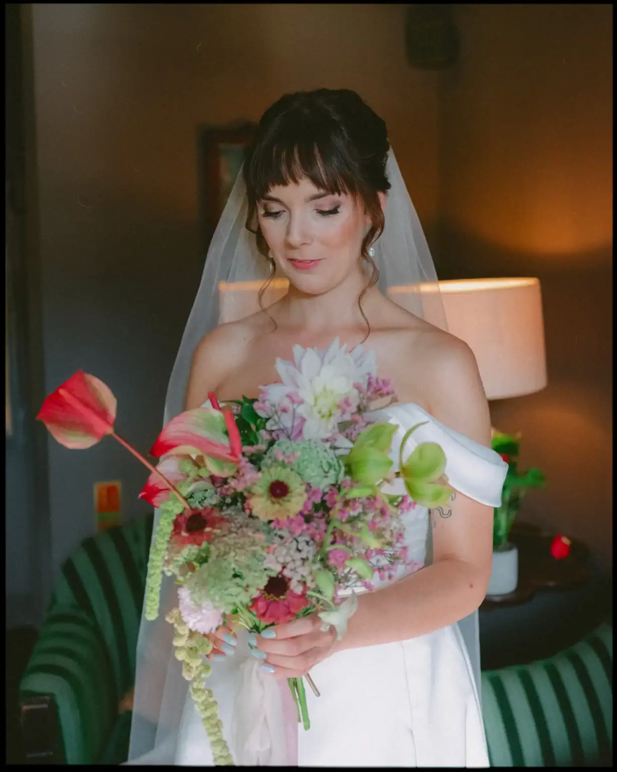 A bride in a white dress and veil holds a colorful bouquet, looking down and smiling softly in a warmly lit room with green-striped furniture—captured in vibrant, colourful wedding photography by a Rye wedding photographer.