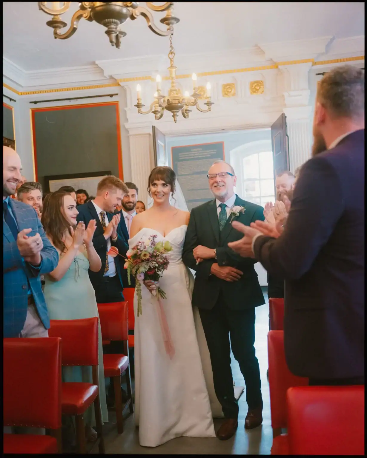 A bride in a white dress, holding a bouquet, walks down the aisle arm-in-arm with an older man. Guests in formal attire smile and applaud as they pass red chairs—captured in candid wedding photography Rye beneath a sparkling chandelier.