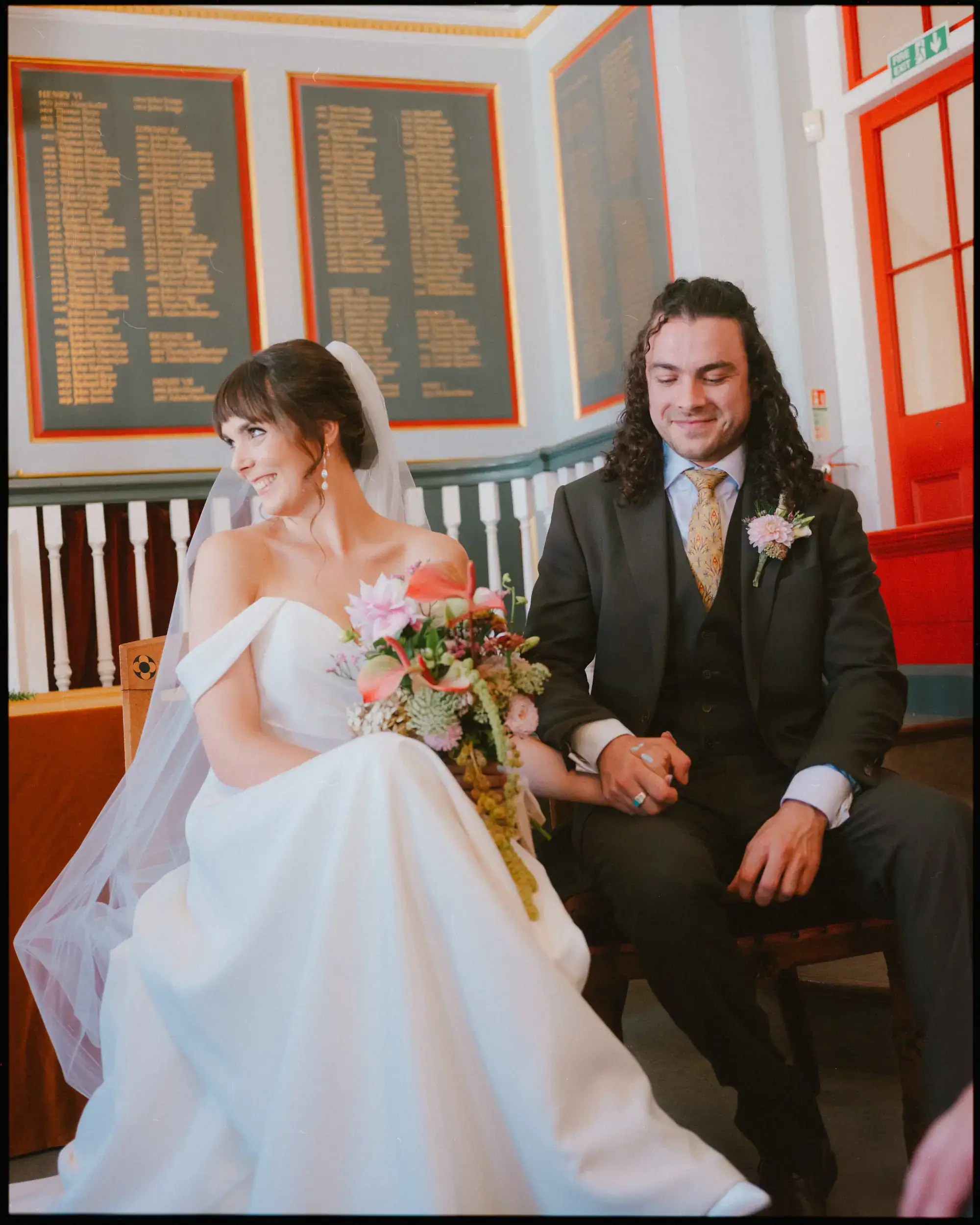 A bride in a white dress and veil sits holding a bouquet, smiling at her groom. The groom, in a dark suit with a floral boutonnière, sits beside her, holding her hand. This moment of colourful wedding photography is set indoors with red and gold decor.