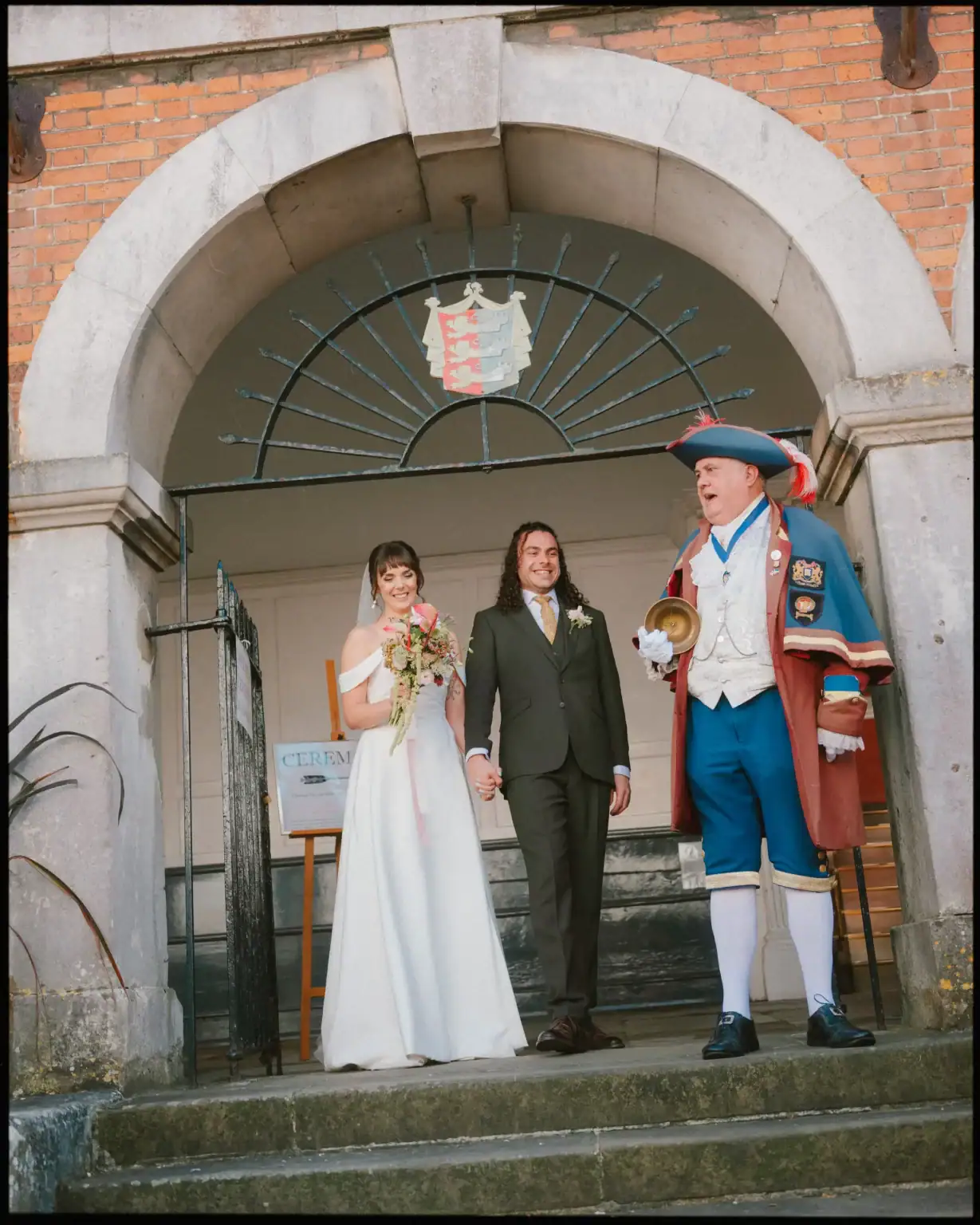 A bride and groom stand hand in hand under a stone archway, smiling. A town crier in a colorful uniform and tricorn hat stands beside them, announcing. The scene captures the charm of an East Sussex wedding venue. A Ceremony sign is visible in the background.
