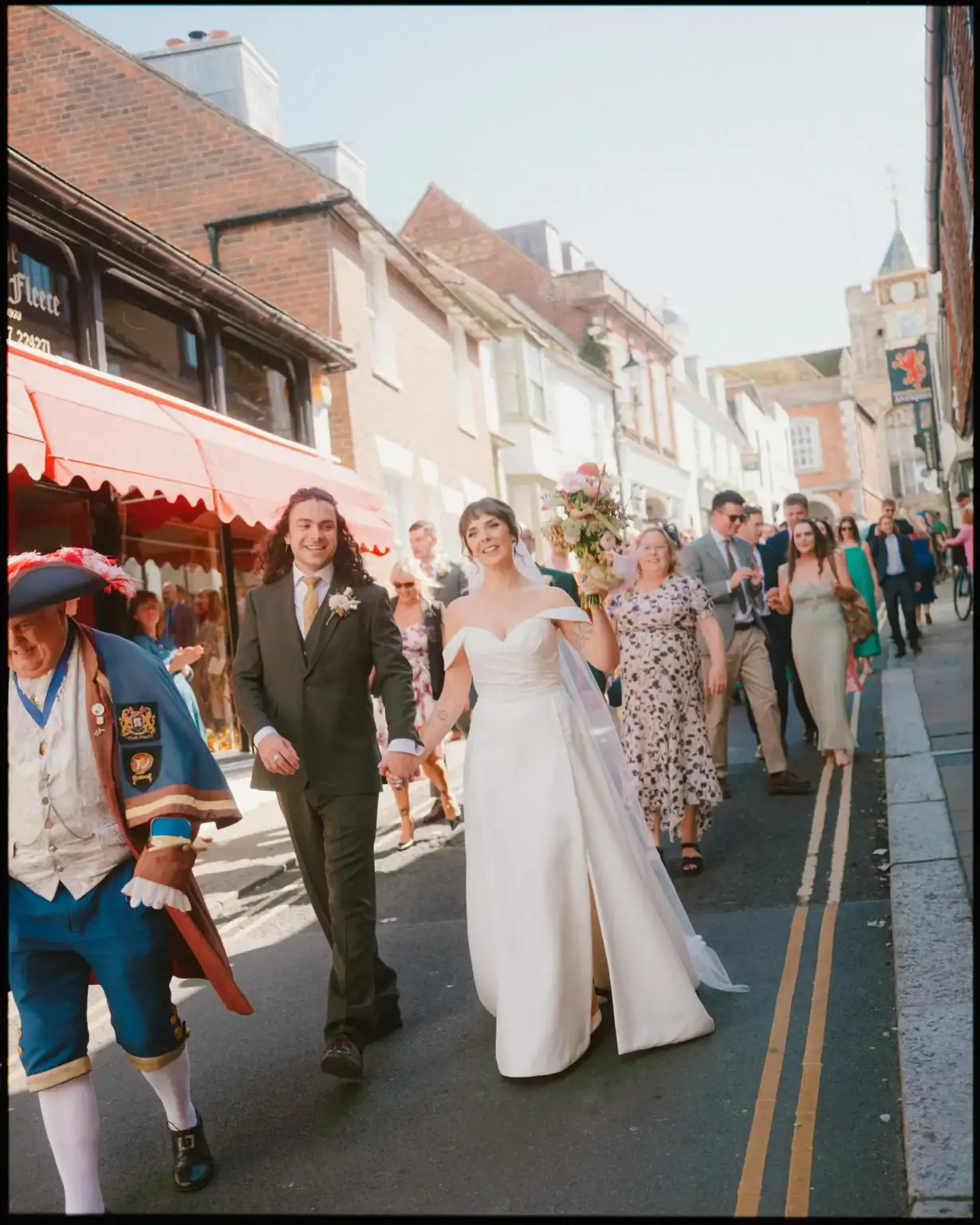 A bride and groom walk hand in hand down a sunny street in Rye, smiling, followed by well-dressed guests. The candid wedding photography captures the moment beautifully as they pass the George in rye and charming buildings.
