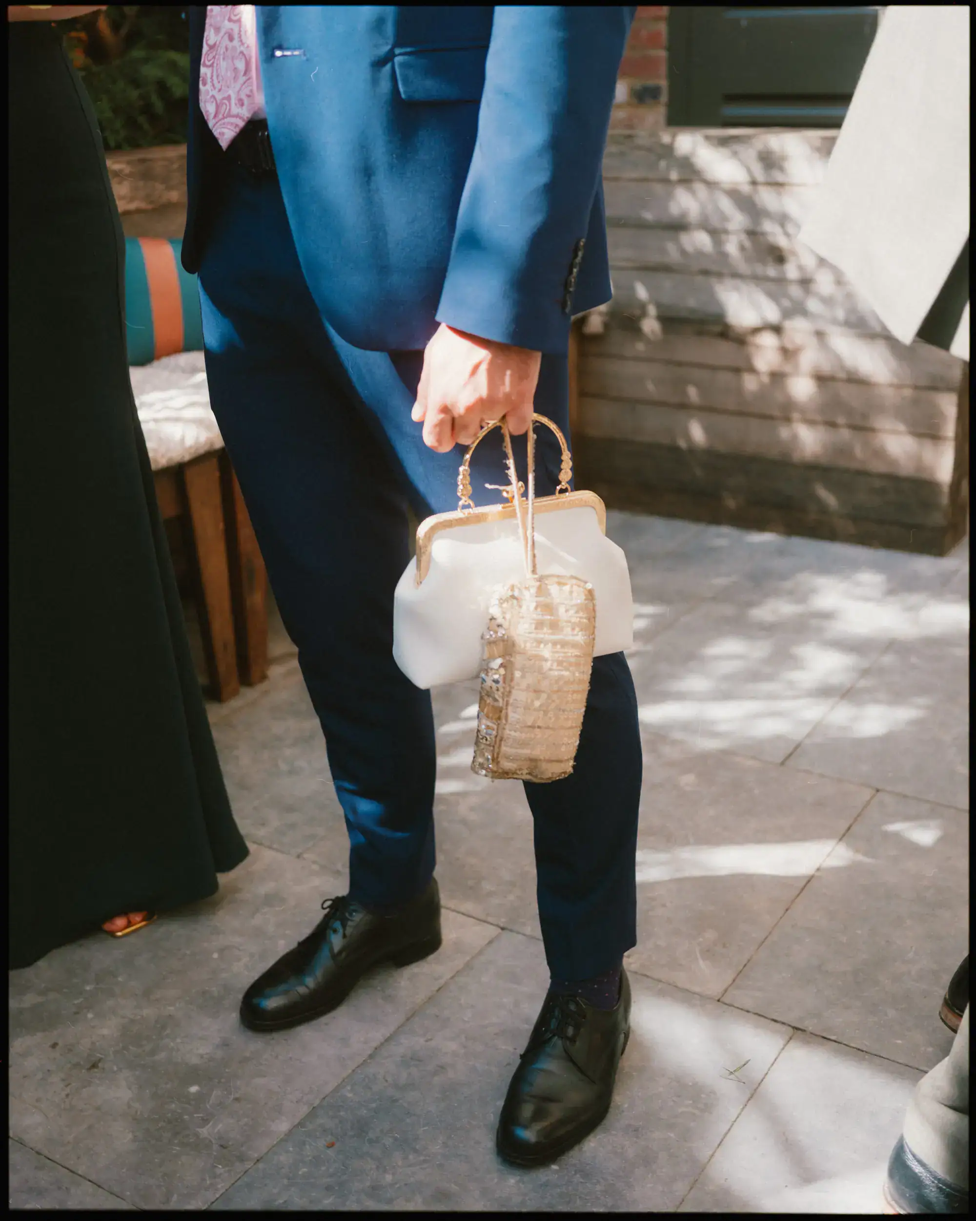 A person in a navy blue suit stands outdoors on a stone patio, holding two handbags—one white and one gold. Captured in candid wedding photography style, sunlight filters through the scene, with a wooden bench in the background.