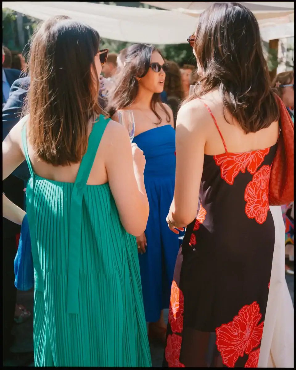 Three women in colourful summer dresses chat with drinks, seen from behind, at a sunny outdoor wedding drinks reception