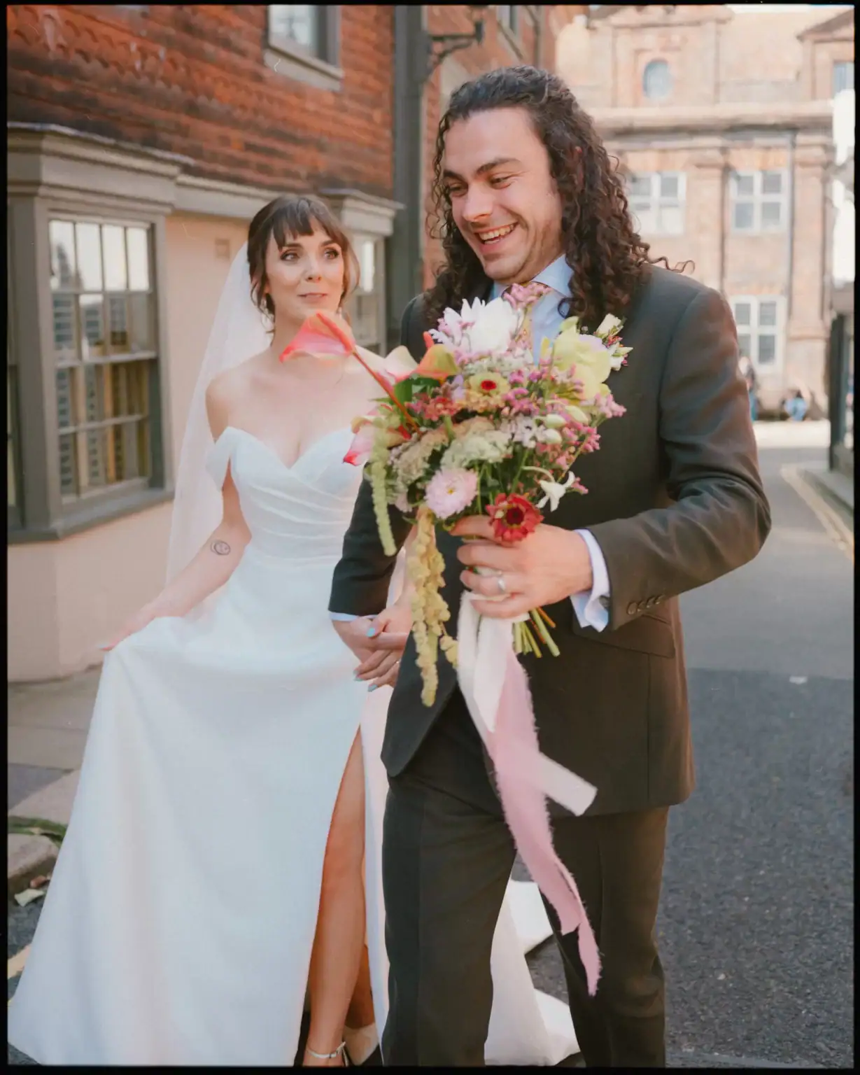 A bride in a white dress with a thigh-high slit smiles at her groom holding a colorful bouquet as they walk outdoors—perfect candid wedding photography Rye at the beautiful George Hotel Rye wedding venue.
