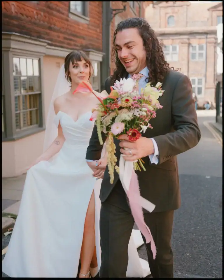 A bride in a white dress with a thigh-high slit smiles at her groom holding a colorful bouquet as they walk outdoors—perfect candid wedding photography Rye at the beautiful George Hotel Rye wedding venue.