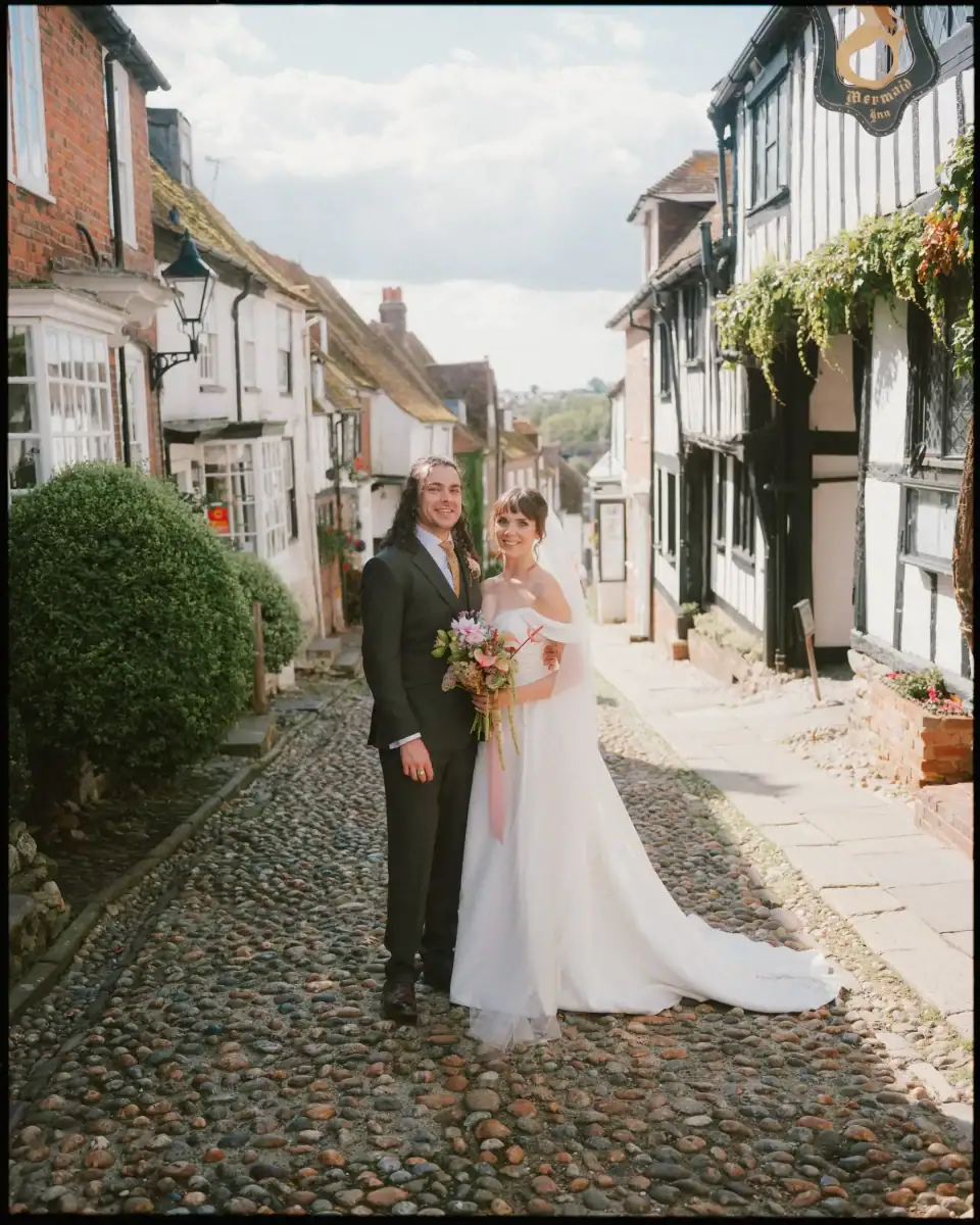 A bride and groom stand together, smiling, on Mermaid Street in Rye, lined with historic buildings and greenery on a sunny day. Captured by a Rye wedding photographer, the scene showcases the charm of an East Sussex wedding venue.