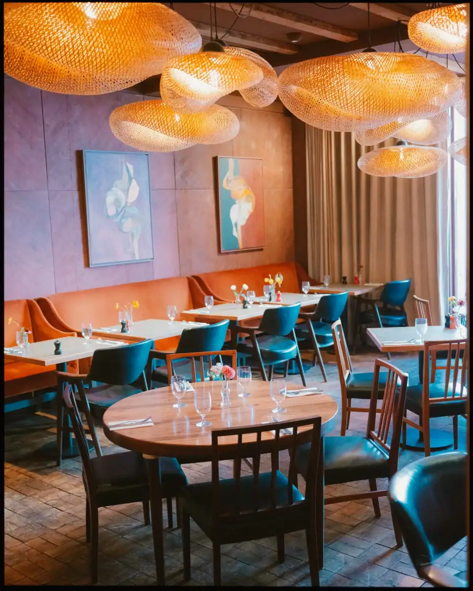 A stylish restaurant interior with modern chairs, warm lighting from woven pendant lamps, and abstract art—perfect for colourful wedding photography or capturing elegant George Hotel Rye wedding photos amid beautifully set tables and large curtained windows.