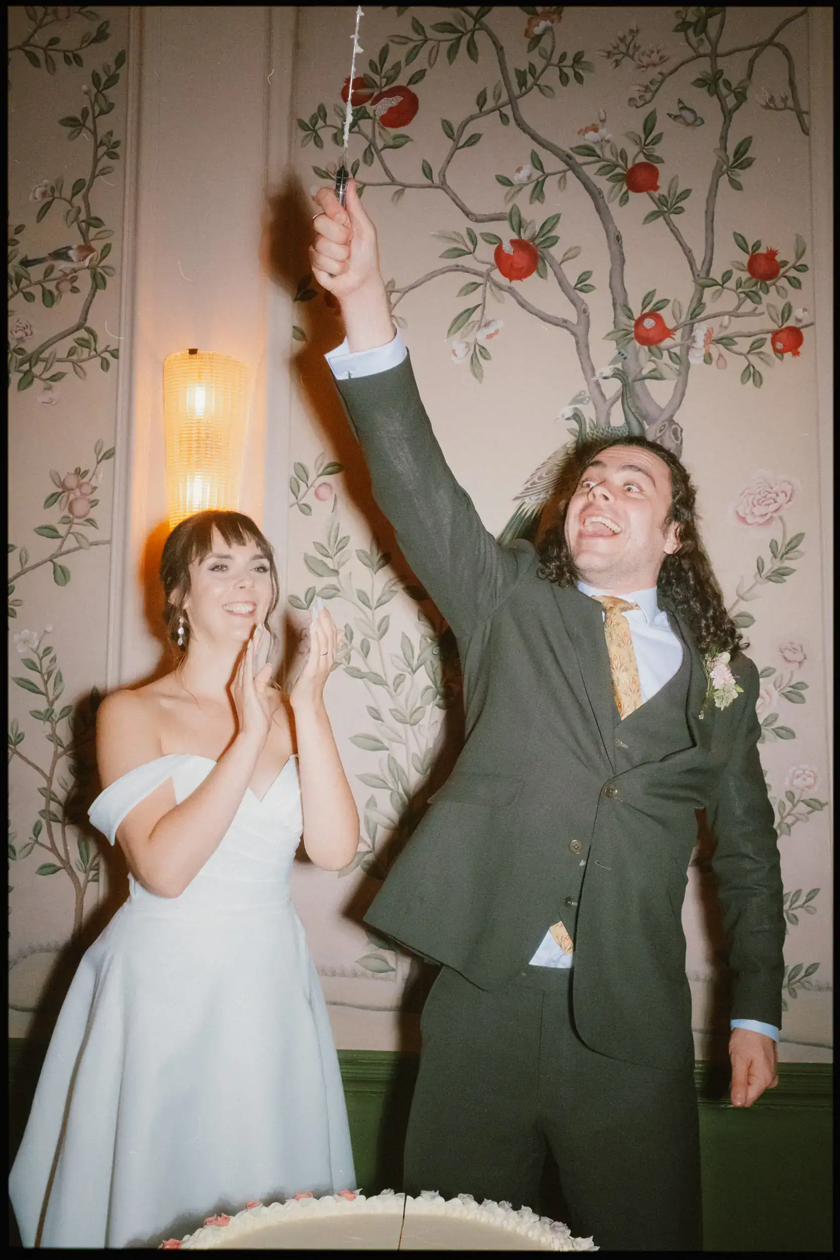 A bride in an off-shoulder white dress smiles and claps as a groom in a suit excitedly raises a cake knife. Captured in vibrant style, this George in Rye wedding photo features a floral-patterned wall and pure joy.