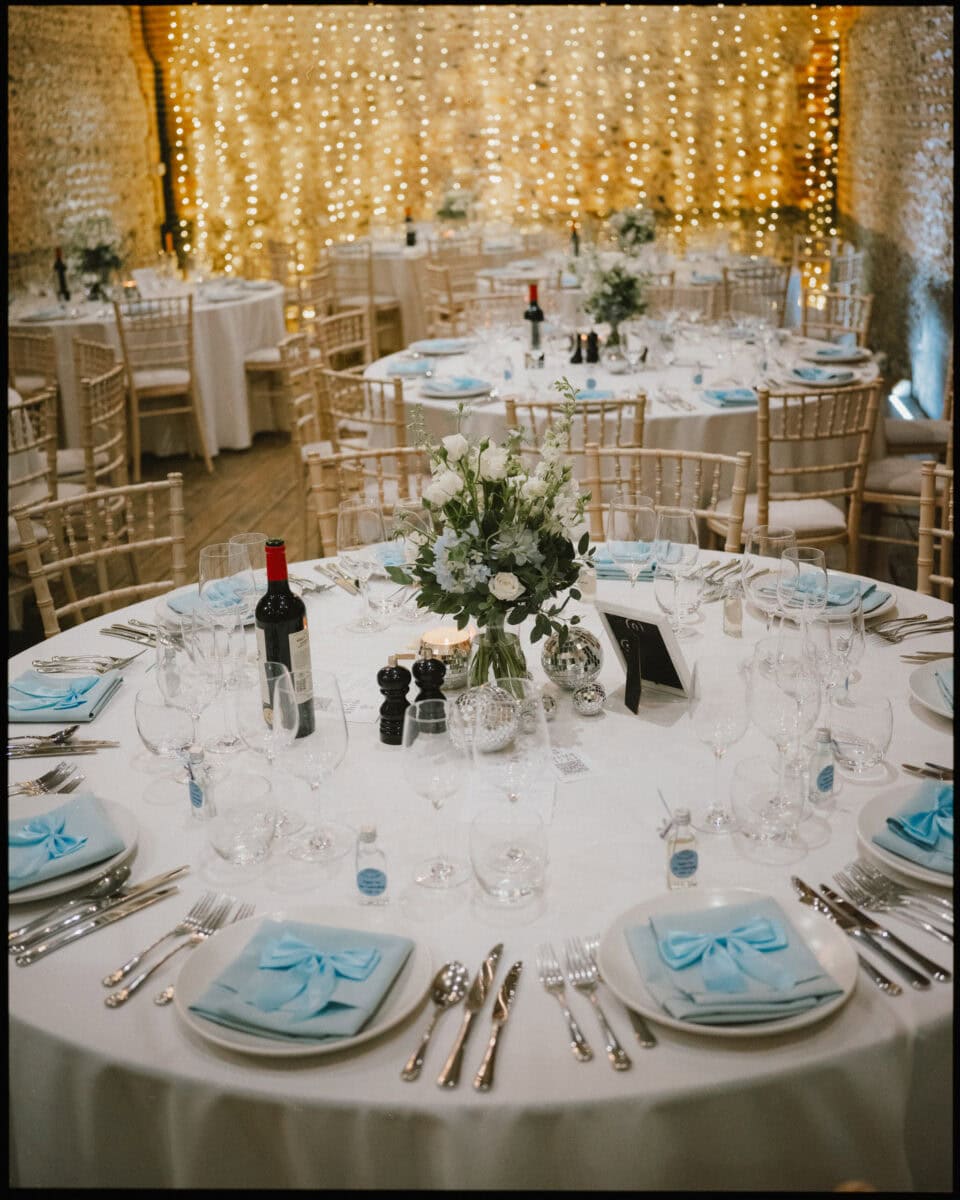 Wedding breakfast tables with blue napkin bows, floral centrepieces, and fairy lights at Cissbury Barns in West Sussex.