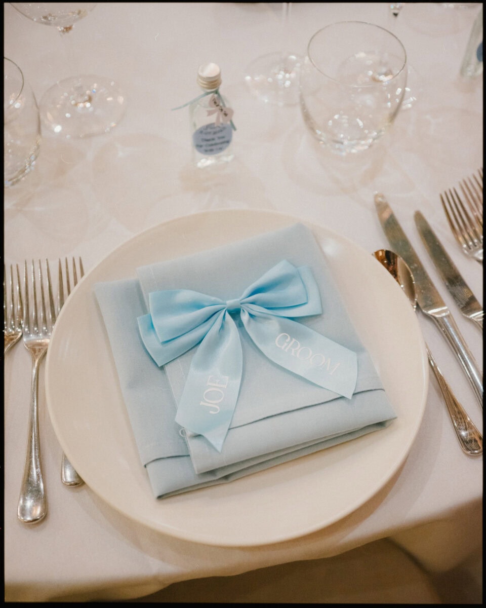 Wedding place setting with blue napkin, personalised ribbon bow, and mini bottle favour at Cissbury Barns in West Sussex.