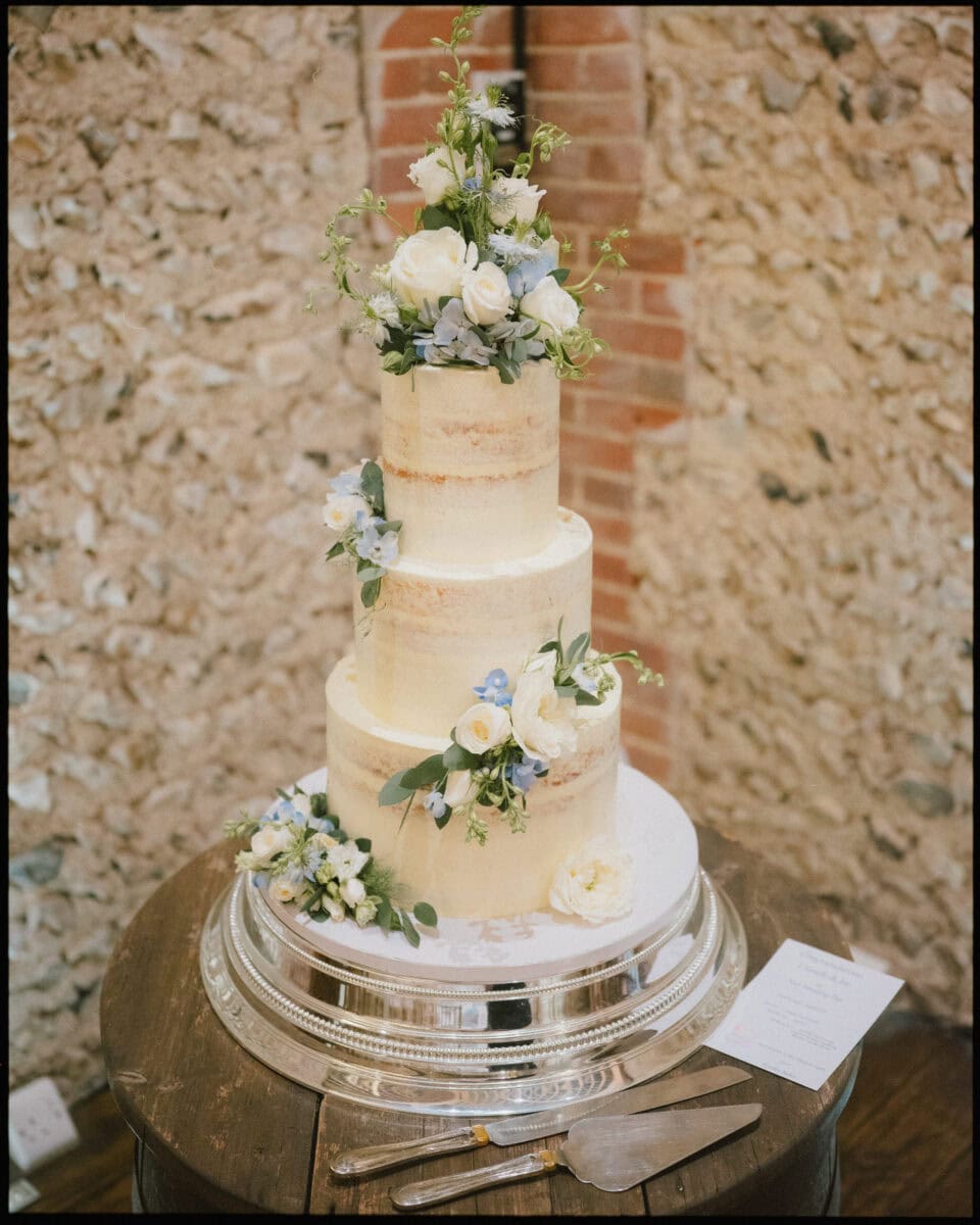 Three-tier semi-naked wedding cake with white and blue flowers on a silver stand at Cissbury Barns in West Sussex.