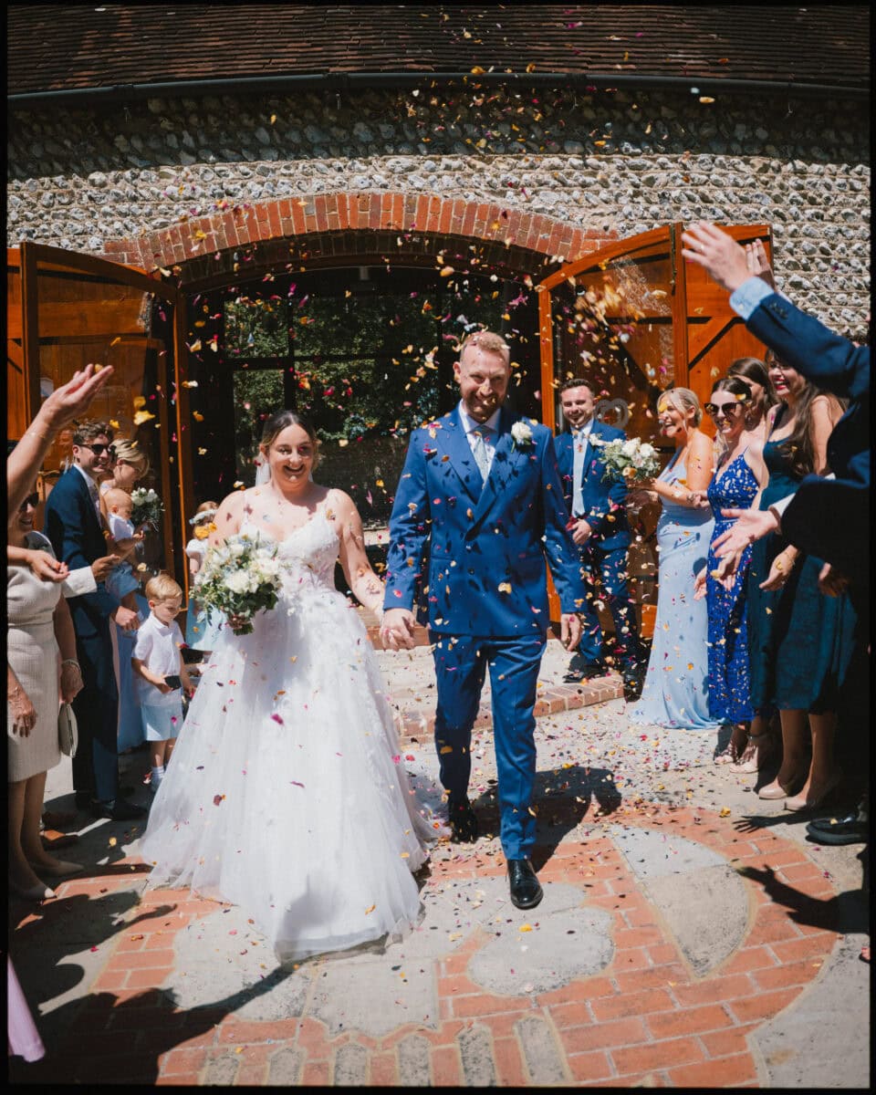 Bride and groom walking through confetti toss outside Cissbury Barns in Findon, West Sussex, surrounded by wedding guests.