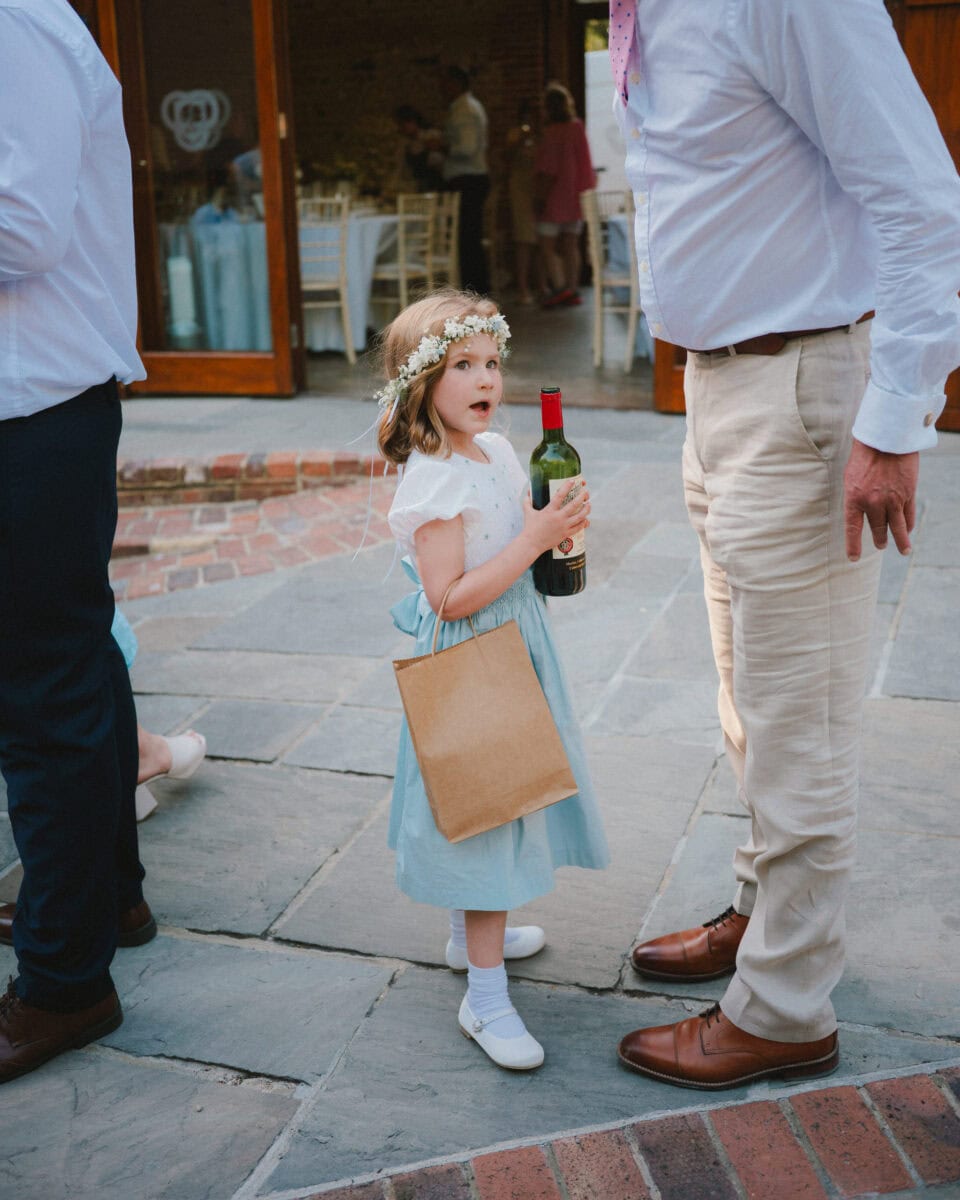 Flower girl wearing a blue dress and flower crown holding a wine bottle during a candid moment at Cissbury Barns wedding in West Sussex.