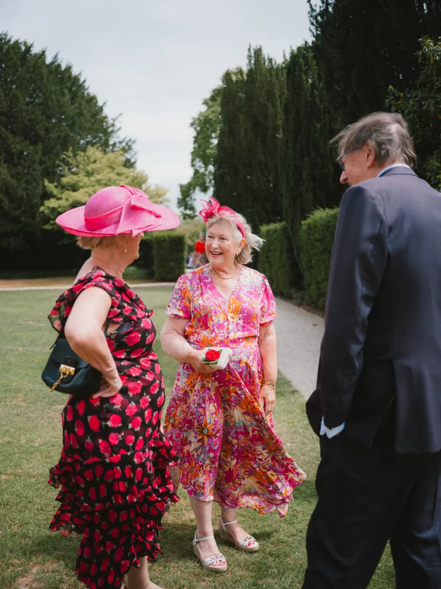 Three people stand outdoors on grass, chatting and smiling. Two women wear bright floral dresses and large pink hats, while a man in a dark suit faces them. Tall trees and hedges line the background.