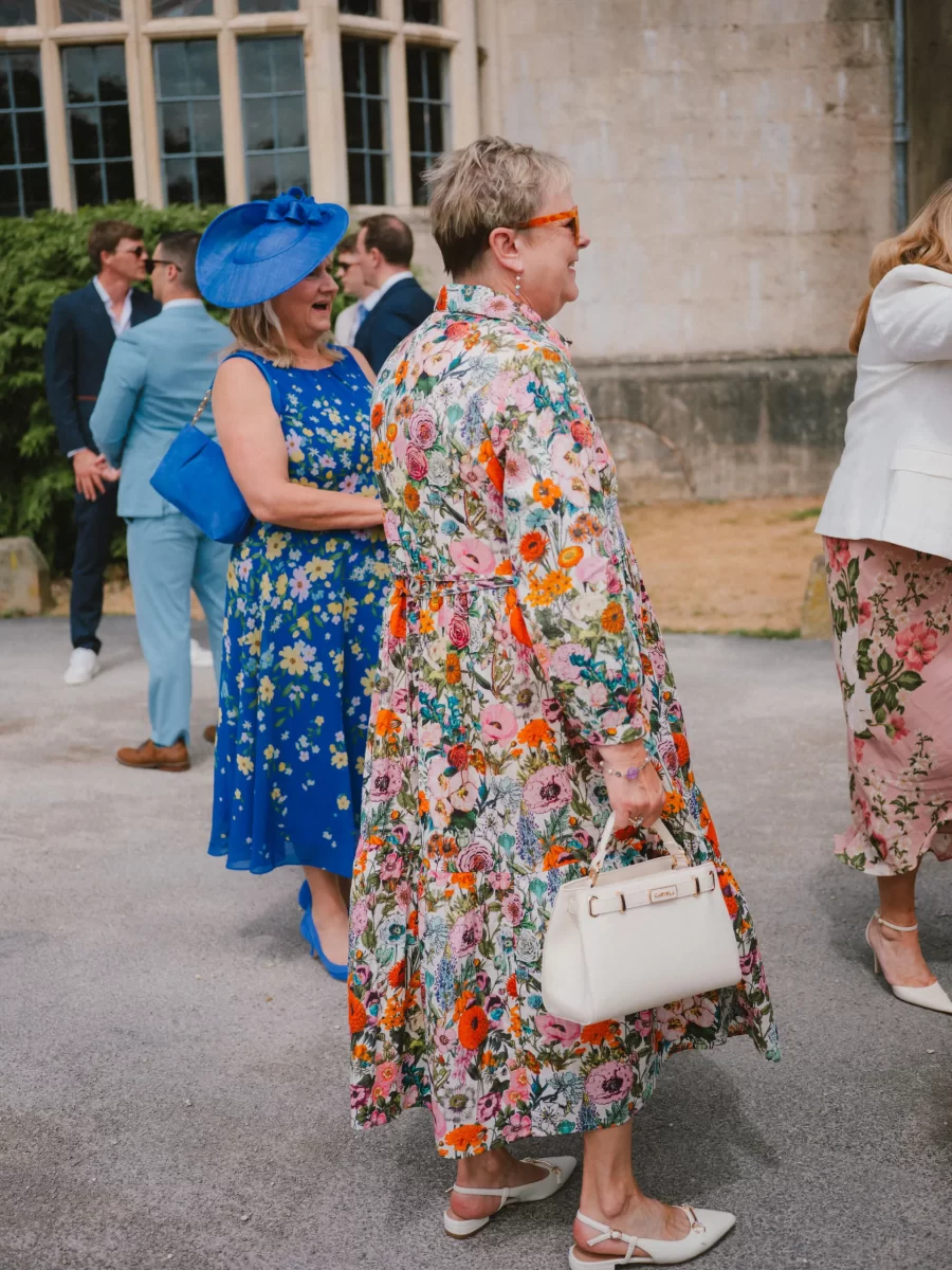 Two women stand outdoors at a formal event, both wearing colorful floral dresses. One also wears a blue hat, the other orange sunglasses and holds a white handbag. Other guests in suits are in the background.