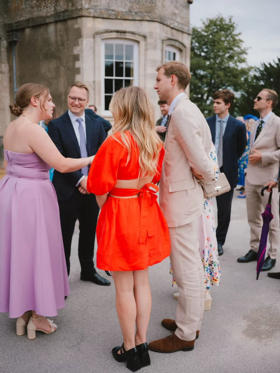 A group of well-dressed people stand and chat outside a stone building. One woman wears a lilac dress, another a bright red dress, and a man in a light suit stands beside them. Others in suits are in the background.