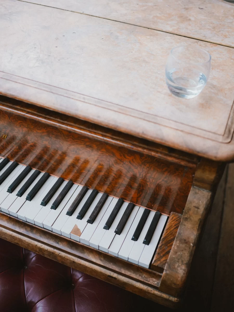 A close-up of part of a wooden piano keyboard with a glass of water resting on its closed top lid. The piano has a vintage, polished appearance and is next to a maroon tufted leather seat.