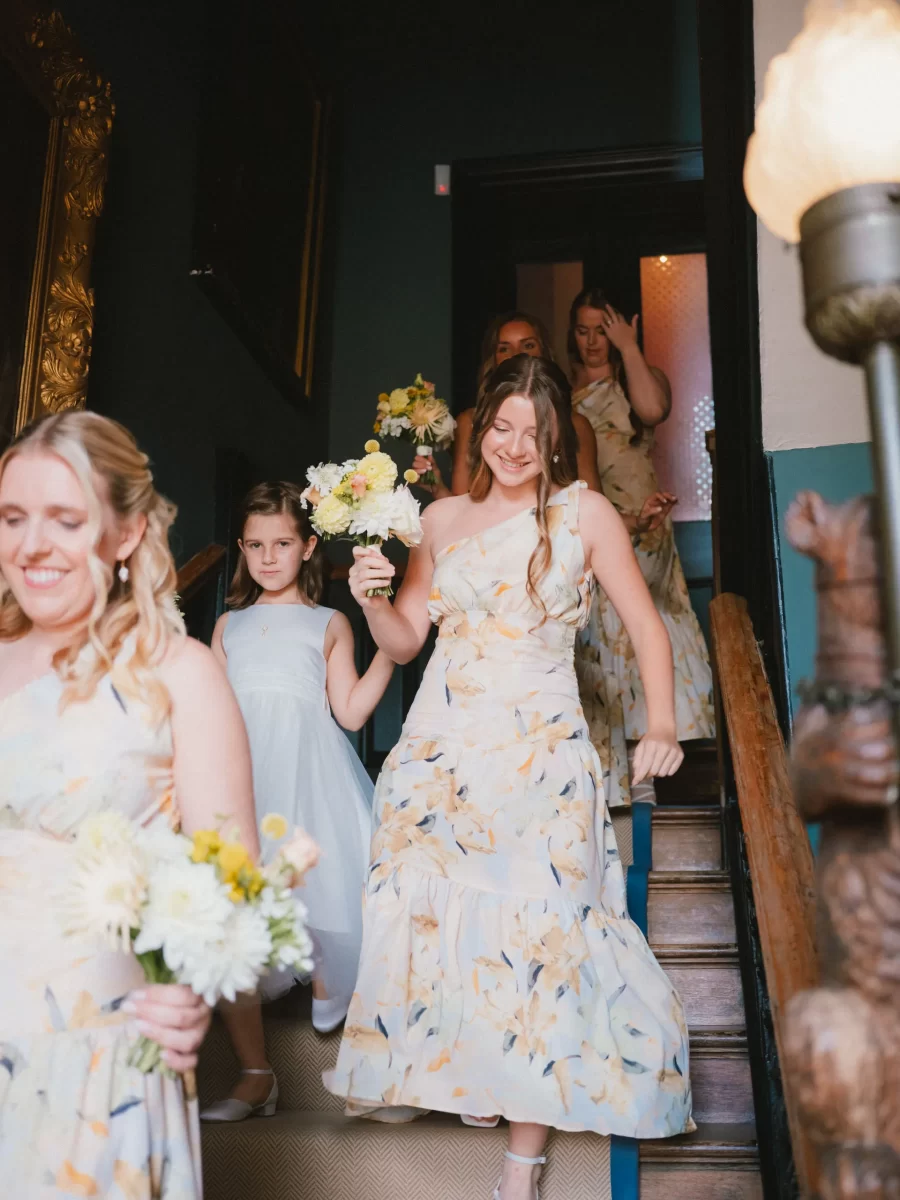A group of bridesmaids in floral dresses and a young girl in a white dress walk down a staircase, smiling and holding bouquets of flowers. The setting appears to be indoors with warm lighting.
