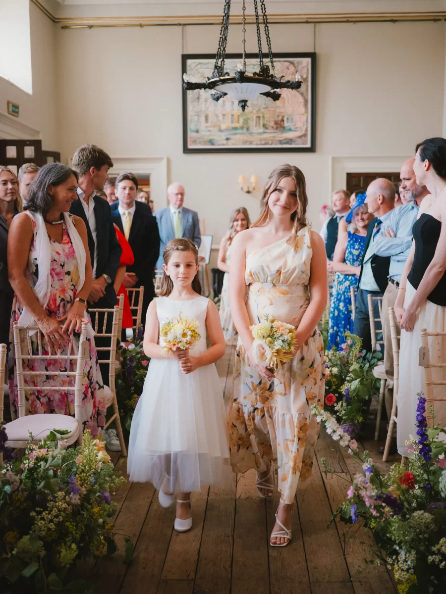 Two girls in floral dresses walk down a flower-lined aisle at an indoor wedding ceremony, holding small bouquets. Guests in colorful attire are seated and watching, and a painting hangs on the wall behind them.