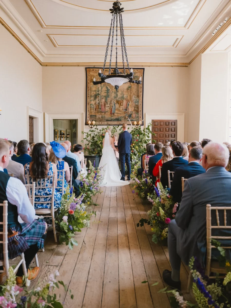 A bride and groom stand at the altar in a light-filled room at Elmore Court, exchanging vows before seated guests. The aisle is lined with chairs and colorful flower arrangements, and a large tapestry hangs on the wall behind them.