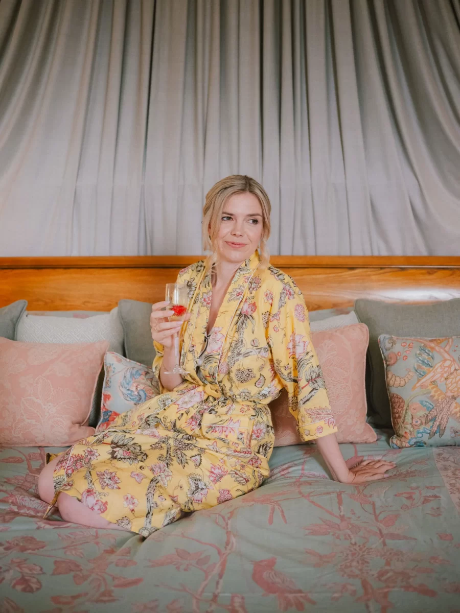 A bridein a yellow floral robe sits on a bed with pastel pillows, holding a drink in her hand and looking to the side. The background features draped curtains at Elmore Court