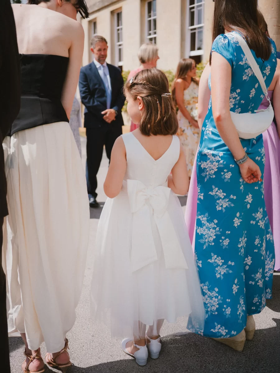 A young girl in a white dress with a large bow stands outside between two women, one in a white skirt and the other in a blue floral dress, with people and a building visible in the background.
