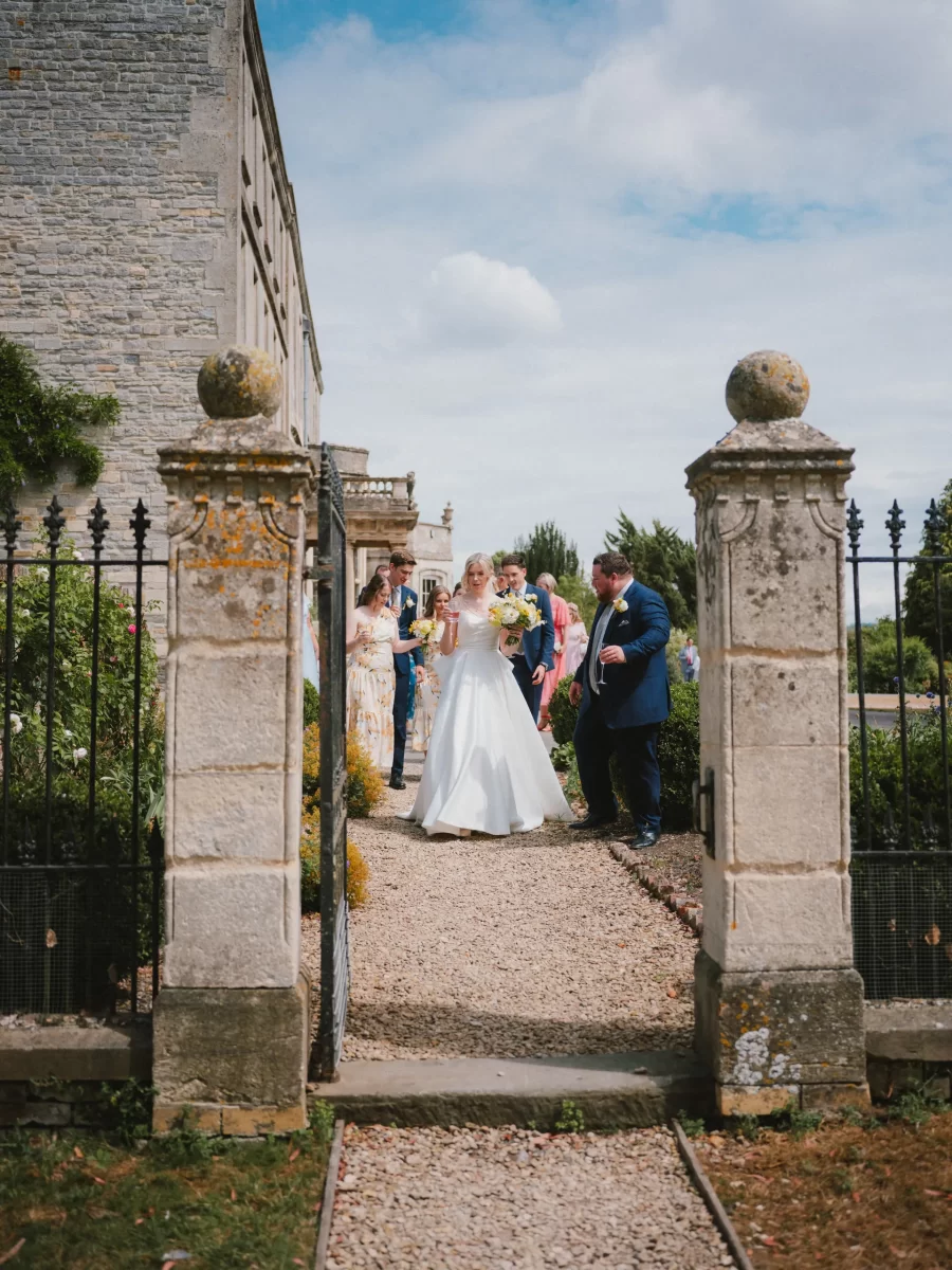 A bride in a white gown walks down a gravel path outside Elmore Court, through a stone gate, followed by bridesmaids and guests, with an old stone building and greenery in the background on a partly cloudy day.