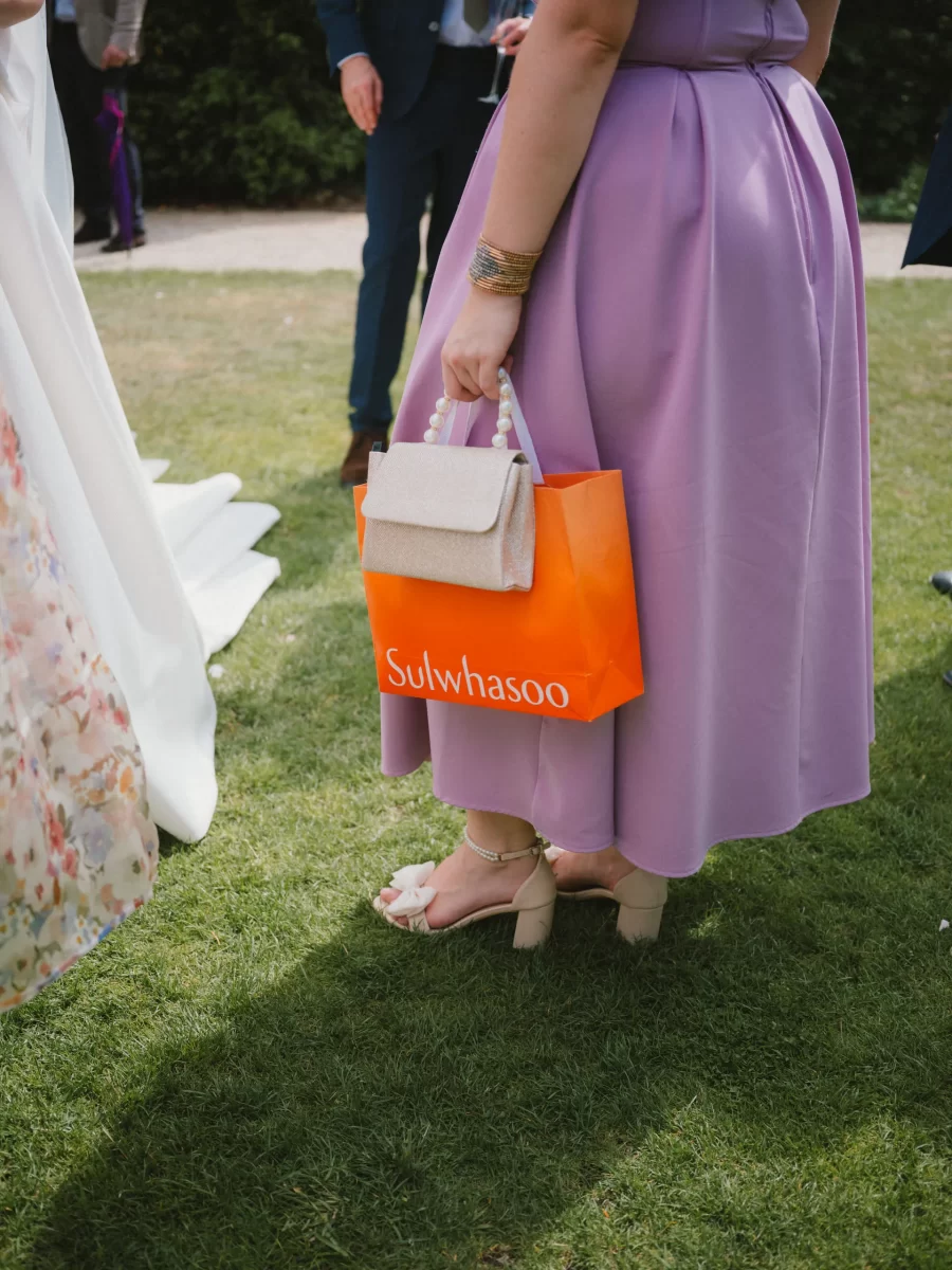 A woman in a lavender dress holds an orange Sulwhasoo shopping bag and a small beige clutch, standing on grass in heeled sandals at an outdoor event. Other people in formal attire are partially visible around her.