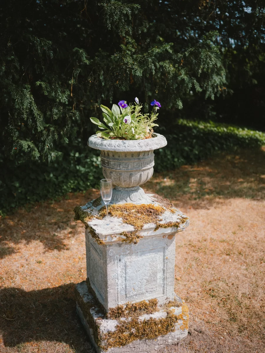 A stone pedestal with patches of moss supports a decorative planter filled with purple and white flowers. An empty champagne glass rests on the pedestal. The scene is outdoors, with grass and trees in the background.