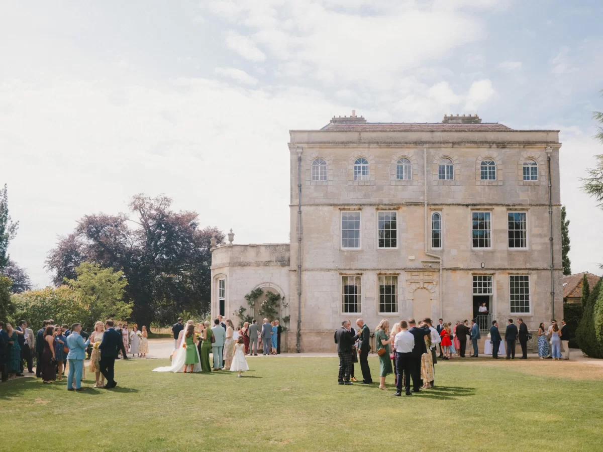 A group of people in formal attire gather on the lawn in front of a large, elegant stone mansion under a partly cloudy sky, suggesting a celebratory outdoor event.